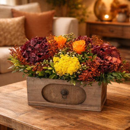 Centro de flores preservadas con rosas naranjas y hortensias burdeos en caja de madera vintage sobre mesa rústica.