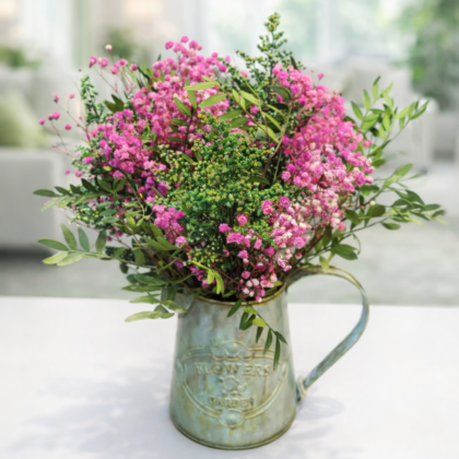 Regadera metálica vintage con paniculata rosa y follaje verde sobre mesa blanca.